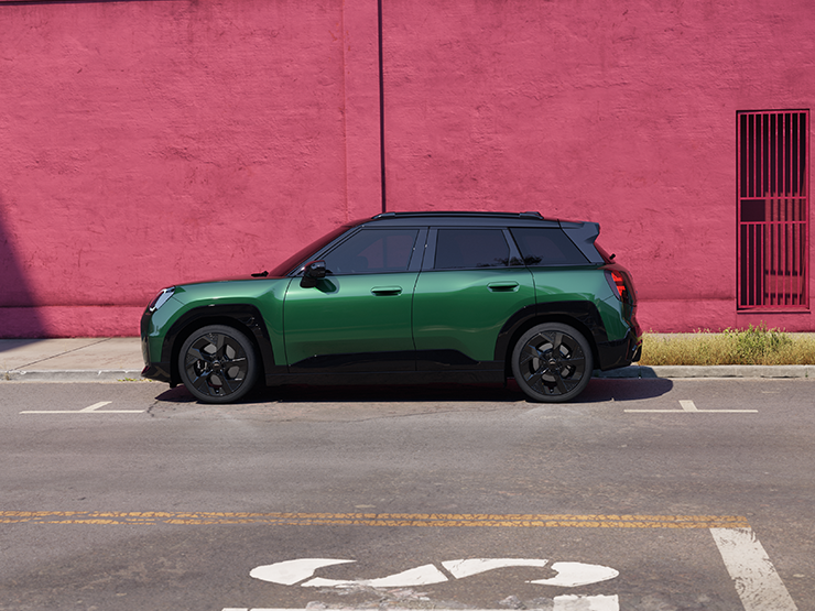 Side view of a MINI Aceman in British Racing Green IV Metallic parked on a street with a pink wall in the background.