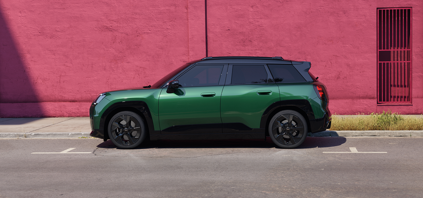 Side view of a MINI Aceman in British Racing Green IV Metallic parked on a street with a pink wall in the background.