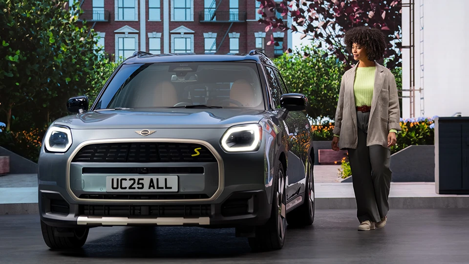 Front view of a MINI Countryman in Slate Grey Metallic with a person walking beside it in an urban setting.