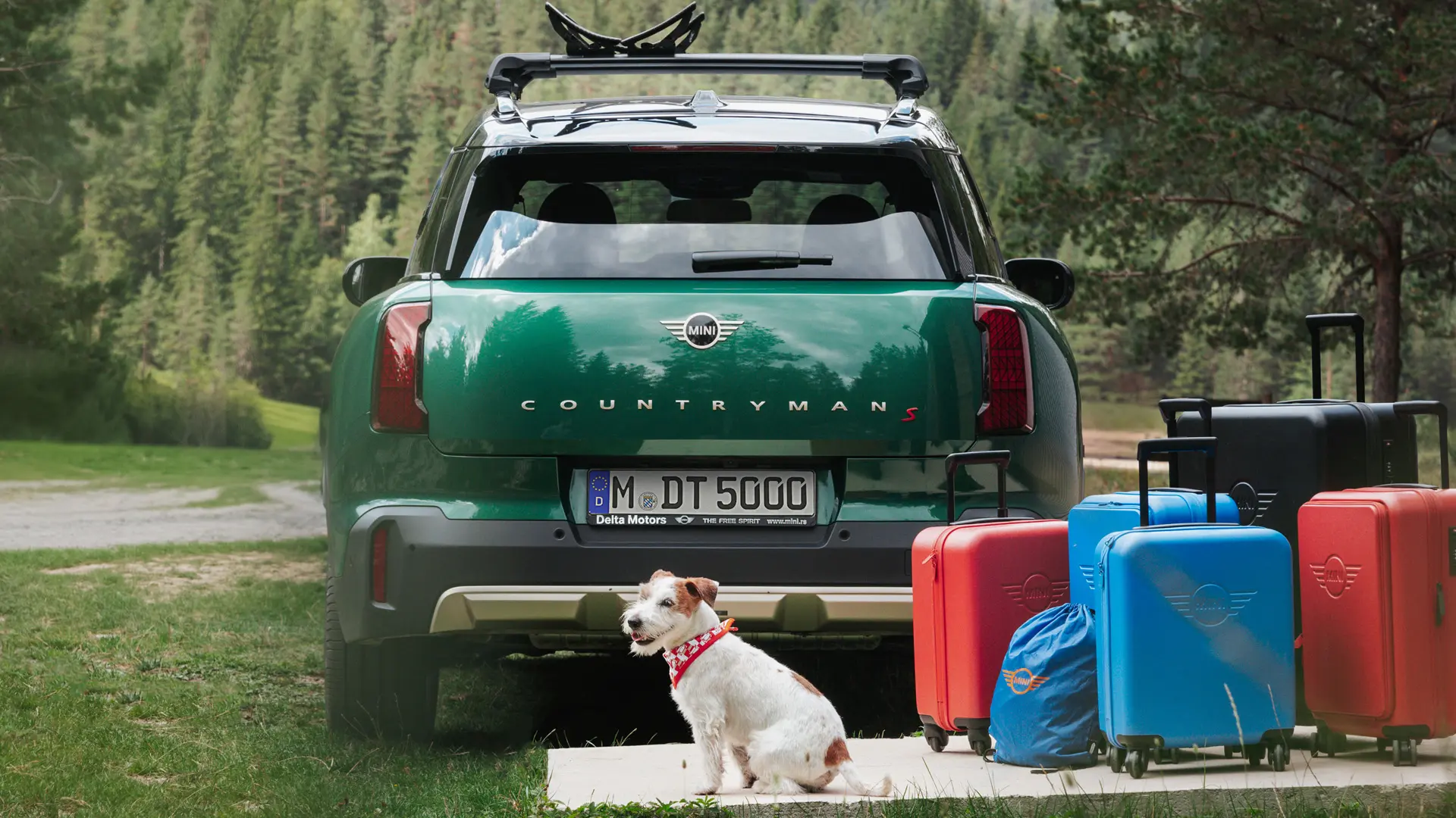 Grænn MINI Countryman með ýmsum farangurshlutum í boði frá MINI Lifestyle versluninni sem stendur fyrir aftan bifreiðina ásamt Jack Russell hundi. 